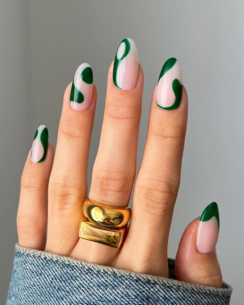 Zdjęcie dłoni ze zbliżeniem na długie, migdałowe paznokcie ze wzorem swirl nails w kolorze zielonym na beżowej bazie. Na środkowym palcu widać dwa masywne złote pierścionki.