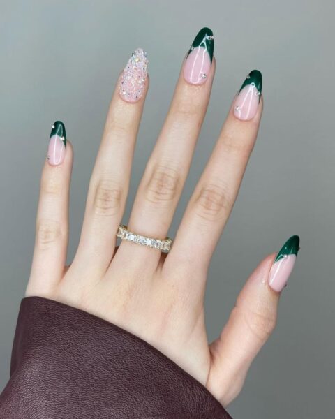 Zdjęcie dłoni ze zbliżeniem na długie, migdałowe paznokcie ze wzorem french manicure z końcówkami w kolorze butelkowej zieleni, oraz ozdobione kryształkami. Paznokieć palca środkowego cały jest ozdobiony kryształkami. Na palcu środkowym widać pierścionek z brylantami. Widać fragment rękawa skórzanej, bordowej kurtki.
