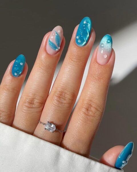 Zdjęcie dłoni z długimi, owalnymi paznokciami w stylu mermaid nails. Są pomalowane lśniącym niebieskim kolorem przypominającym morską wodę, widać też wzór fragmentu plaży, perełki oraz wzór płetw syreny. Na środkowym palcu widać srebrny pierścionek z kamieniem.