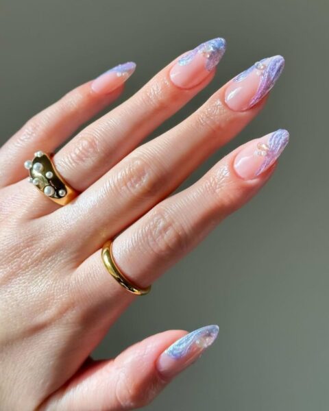 Zdjęcie dłoni ze zbliżeniem na paznokcie mermaid nails. Paznokcie są długie, mają migdałowy kształt. Są pomalowane na kolor fioletowy w stylu french manicure, strukturalnym wzorem swirl przy brzegach i perełkami 3D. na palcach widać złote pierścionki.