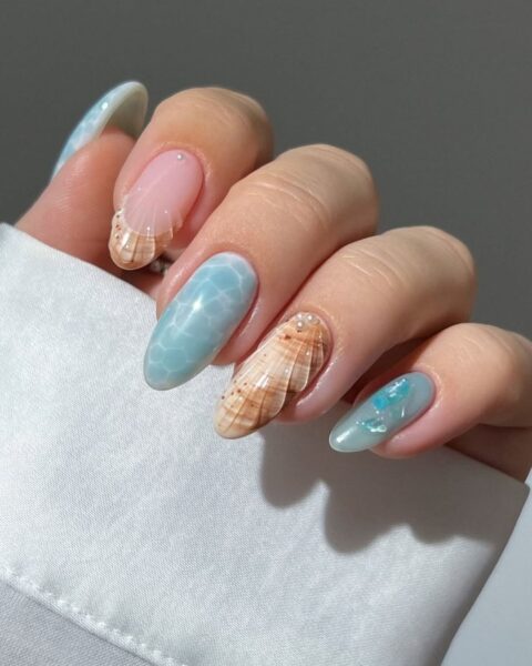 Zdjęcie dłoni ze zbliżeniem na paznokcie długie, migdałowe paznokcie ze wzorem mermaid nails. Dwa paznokcie są ozdiobone brązowo-beżowym wzorem muszli oraz wzorem strukturalnym muszli. Pozostałe paznokcie są pomalowane na niebiesko imitujące morską toń. Widać też małe perełki 3D.