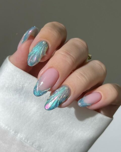Zdjęcie dłoni z długimi, migdałowymi paznokciami w stylu syrenkowym. Są zielone, opalizujące mermaid nails z muszlami 3D, strukturalnym wzorem muszli oraz opalowym różowym blaskiem. Widać fragment białego rękawa koszuli.