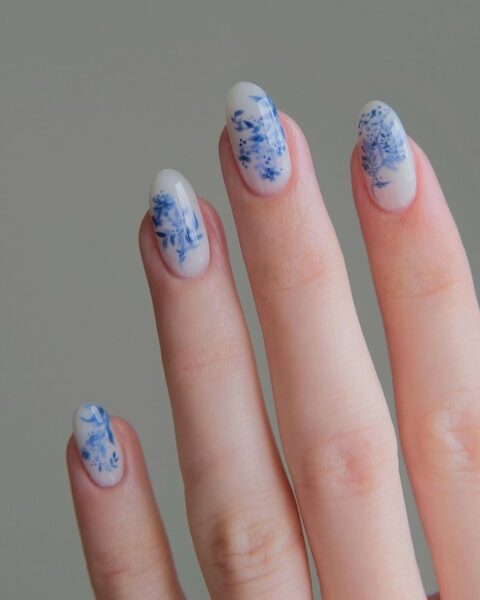 Zdjęcie dłoni ze zbliżeniem migdałowe paznokcie pomalowane na kolor mlecznej bieli i ozdobione niebieskim wzorem kwiatów i gałęzi w stylu nail art.
