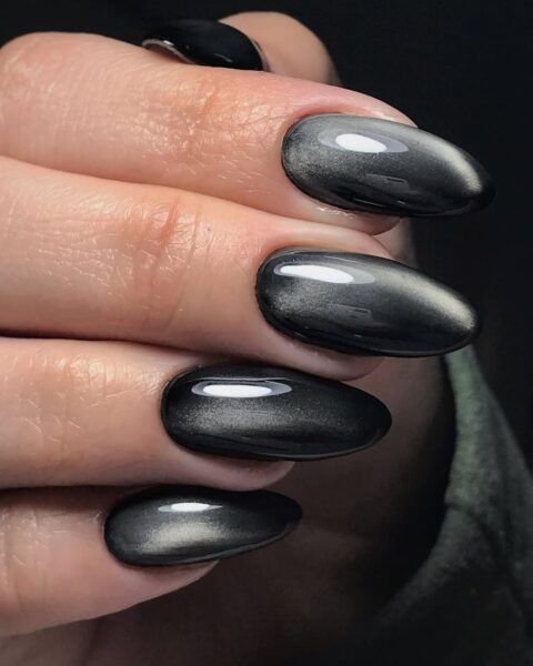 Zdjęcie dłoni z długimi, migdałowymi paznokciami pomalowanymi na styl mirror manicure w kolorze czarnym z efektem lustra.