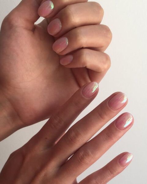 Zdjęcie dłoni z krótkimi, owalnymi paznokciami w kolorze nude i efektem mirror nails z opalowym efektem z zielonym poblaskiem. Dłonie pozują tak, by prezentować manicure.
