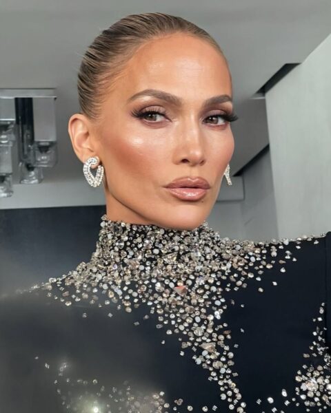 Na zdjęciu jest latynoska gwiazda Jennifer Lopez. Ma włosy związane w kok. W uszach ma srebrne kolczyki. Ubrana jest w czarną sukienkę z długimi rękawami i stójką, ozdobioną kryształami.