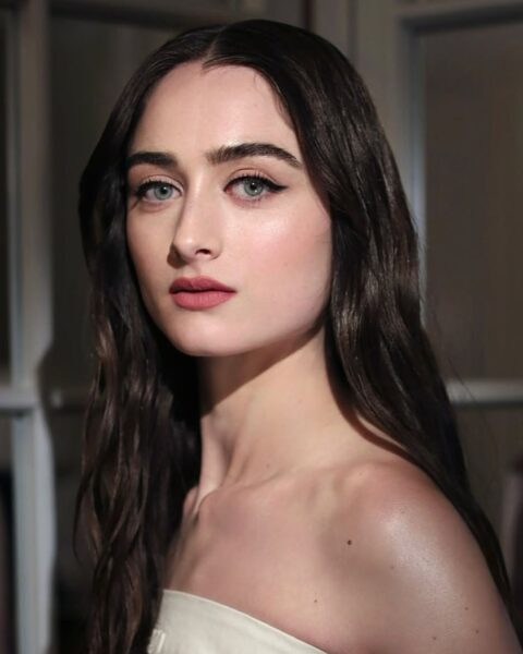 Zdjęcie aktorki Raffey Cassidy. Ma ciemne, długie, rozpuszczone włosy. Ubrana jest w kremowy top bez ramiączek. Ma delikatny makijaż i ciemne brwi zaczesane na bok.