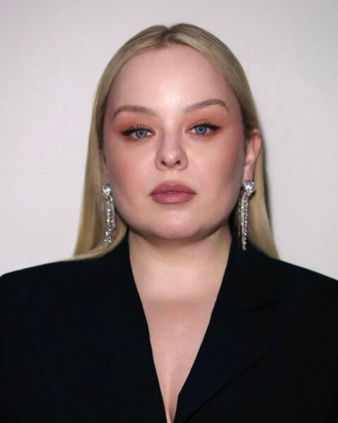Zdjęcie aktorki Nicoli Coughlan. Jest ubrana w czarną marynarkę. Ma w uszach srebrne kolczyki. Włosy blond, proste, rozpuszczone na ramina. Ma delikatny makijaż z modnymi brwiami laminowanymi.