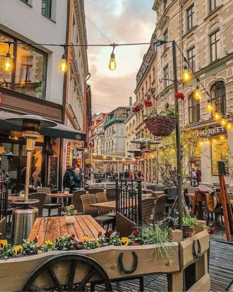 Zdjęcie przedstawiające jedną z ciasnych, otoczonych kamienicami ulicy Rygi, stolicy Łotwy. Widać stoliki restauracyjne jednej z restauracji oświetlonej sznurami lampek.