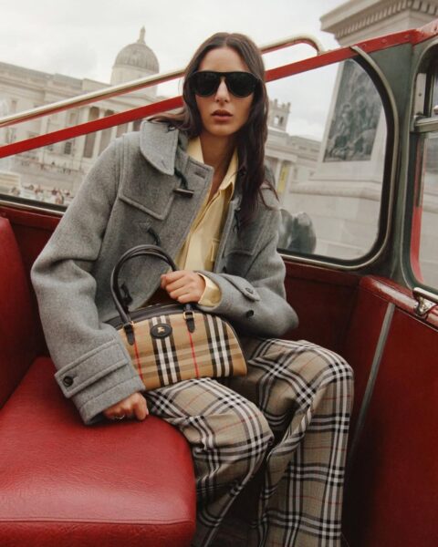 Zdjęcie z kampanii marki Burberry. Widać na nim modelkę w szarym płaszczu, żółtej koszuli oraz szerokich spodniach ze słynną kratką Burberry. Na kolanach trzyma torebkę w tym samym wzorze. Na nosie okulary przeciwsłoneczne. Włosy ciemne, długie, rozpuszczone na ramionach. Modelka siedzi na dachu angielskiego, piętrowego autobusu i jeździ po Londynie.