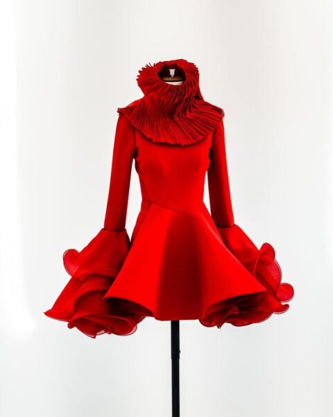 Zdjęcie sukienki marki Alexander McQueen. Sukienka jest na wieszaku. Jest czerwona, ma rozkloszowany dół, długie rękawy z masywnymi, warstwowymi falbankami oraz duży kołnierz typu stójka z zakładowego materiału.
