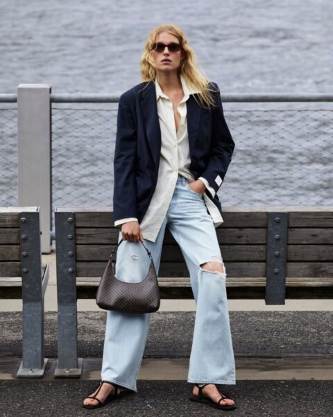 Zdjęcie modelki w stylizacji od marki Calvin Klein. Ma na sobie szerokie jeansy z dziurą na kolanie, długą, białą koszulę, granatową marynarkę, a na nogach czarne sandały na płaskiej podeszwie. W ręce trzyma brązową torebkę. Na nosie okulary przeciwsłoneczne.