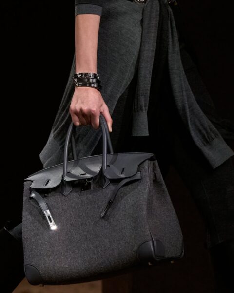 Zdjęcie z pokazu marki Hermès: widać idącą modelkę ubraną na czarną, ale kadr koncentruje się na torebce: jest to szara Birkin Bag.