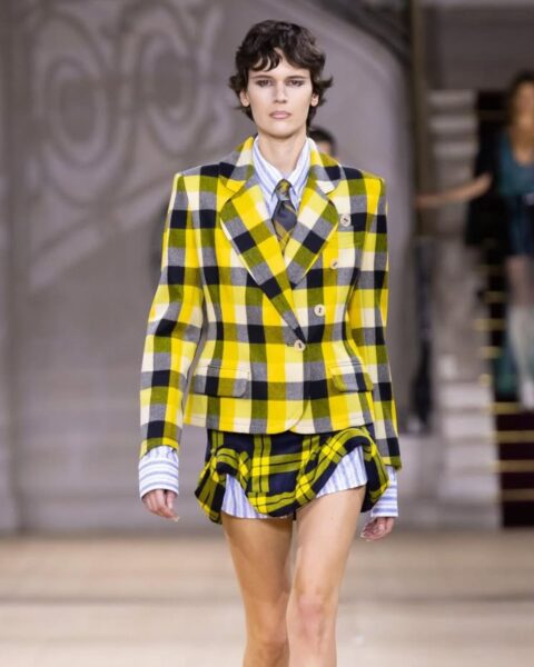 Zdjęcie modelki z pokazu Vivienne Westwood. Ma na sobie żółty komplet w kratkę tartan z marynarką taliowaną oraz mini spódnicą. Ma krótkie, ciemne włosy.