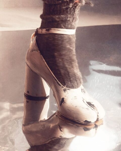 Zdjęcie ze zbliżeniem na nogę i buty marki Maison Margiela. Są to czółenka Tabi w kolorze białym z paskiem wokół kostki i na grubym, masywnym obcasie. Buty są założone na szare, grube skarpetki.