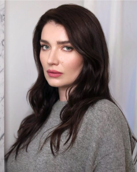 Zdjęcie, na którym jest aktorka Eve Hewson. Ma makijaż nieśmiałej dziewczyny z różowymi policzkami i ustami. Włosy ciemne, długie, rozpuszczone na ramiona. Aktorka jest ubrana w szary sweter. Patrzy prosto w obiektyw.