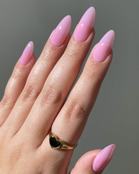 Zdjęcie dłoni ze zbliżeniem na długie paznokcie o migdałowym kształcie w kolorze pink nude jako idealny manicure do biura. Na palcu wskazującym widać złoty pierścionek z motywem serca.