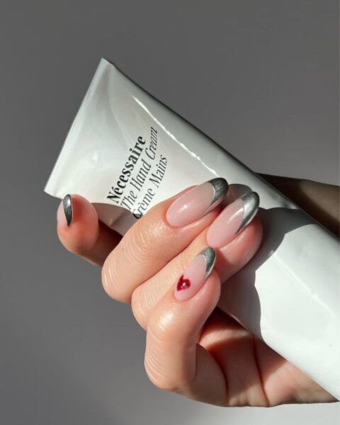 Zdjęcie dłoni ze zbliżeniem na długie, migdałowe paznokcie ze stylizacją french manicure ze srebrnymi końcówkami. Na paznokciu małego palca znajduje się wzór czerwonego serduszka. Dłoń trzyma w ręce tubkę z kremem.