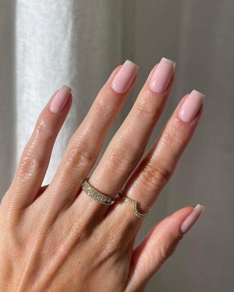 Zdjęcie dłoni ze zbliżeniem na mannequin manicure: długie, kwadratowe paznokcie w kolorze jasnego beżu. Na palcach widać dwa złote pierścionki.