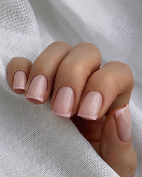 Zdjęcie dłoni ze zbliżeniem na średniej długości, kwadratowe paznokcie ze stylizacją french manicure z różowymi końcówkami. Dłoń pozuje owinięta białym materiałem.