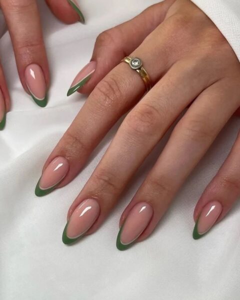 Zdjęcie dłoni ze zbliżęniem długie, migdałowe paznokcie ze wzorem french manicure ale zamiast białej końcówki jest zielona. Na palcu wskazującym widać złoty pierścionek. Dłonie pozują na białym materiale.