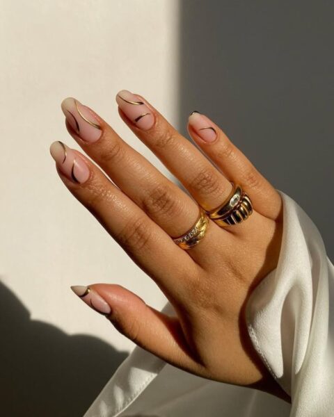 Zdjęcie dłoni ze zbliżęniem długie, migdałowe paznokcie. Są niepomalowane, naked nails, ale ozdobione złotym wzorem swirl. Na palcach widać złote pierścionki. Widać też mankiet rękawa białej koszuli.
