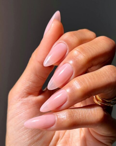 Zdjęcie dłoni z długimi, migdałowymi paznokciami w kolorze różowym w stylu jelly nails. Na palcu widać złoty pierścionek.