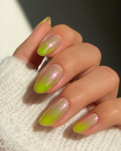 Zdjęcie dłoni z długimi, migdałowymi paznokciami. Są pomalowane na kolor zielony z różowym i pokryte lśniącym pyłkiem w stylu guava nails.