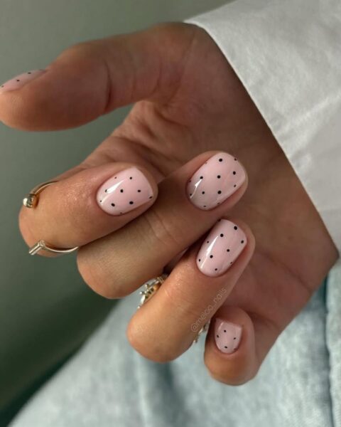 Zdjęcie dłoni z krótkimi, owalnymi paznokciami w kolorze jasnego różu i zdobieniem polka dots, czyli czarnymi kropkami. Na palcach widać złote pierścionki. Widać mankiet białej koszuli.