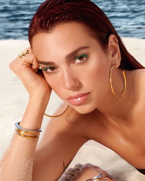 Zdjęcie ze zbliżeniem na twarz piosenkarki Dua Lipa, która pozuje dla kampanii marki YSL Beauty. Ma na sobie mnóstwo biżuterii: kolczyki, bransoletki, pierścionki. Leży na plaży, a za nią widać morze. Ma basenowe usta pomalowane niebieskim błyszczykiem i zielone rzęsy oraz rude, zaczesane na mokro do tyłu włosy.