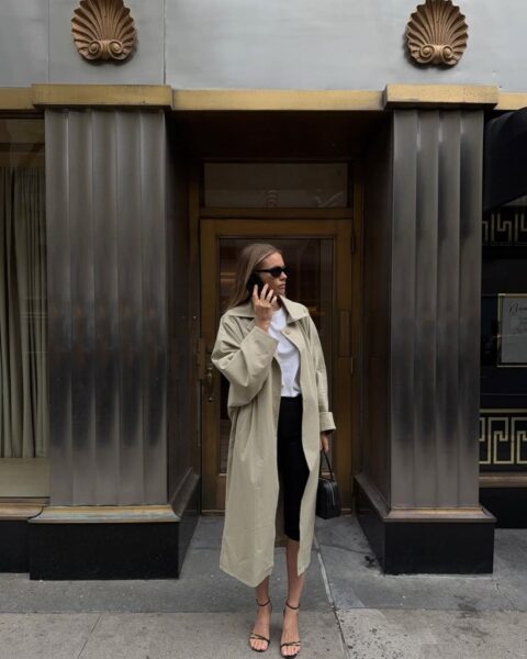 Zdjęcie kobiety, która reprezentuje secret luxury trend: ubrana jest w beżowy trencz, czarne rybaczki, białą bluzkę. Ma w ręce czarną torebkę, na nosie okulary przeciwsłoneczne. Trzyma telefon przy uchu. Włosy blond, rozpuszczone. Stoi przy wejściu do eleganckiego, zabytkowego budynku w stylu art deco.