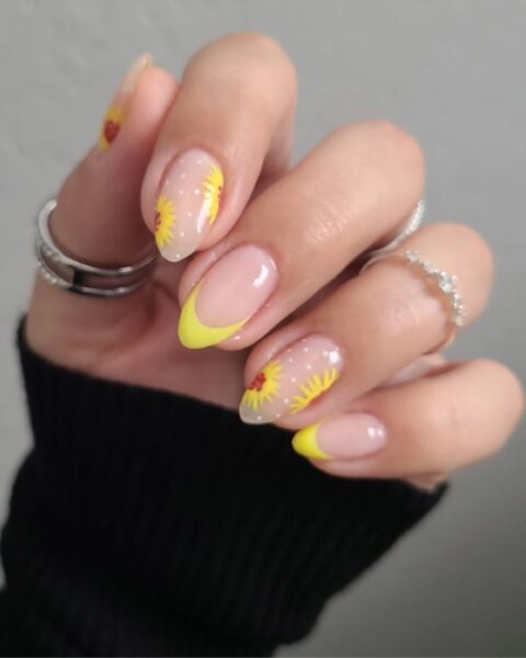 Zdjęcie dłoni ze zbliżeniem na słonecznikowe paznokcie: mają migdałowy kształt, pomalowane w stylu french manicure w kolorze żółtym i ozdobione wzorem słoneczników. Na palcu kciuku widać srebrny pierścionek.