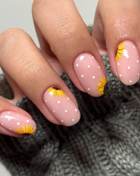 Zdjęcie dłoni ze zbliżeniem na owalne, średniej długości paznokcie w kolorze nude, ozdobione wzorem polka dots (groszki) oraz wzorem słoneczników.