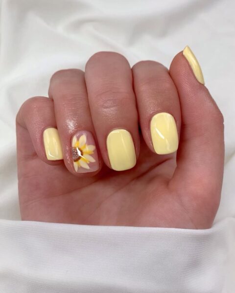 Zdjęcie dłoni ze zbliżeniem na krótkie, kwadratowe paznokcie pomalowane na kolor butter yellow oraz ozdobione wzorem słoneczników. Dłoń pozuje na tle białego materiału.