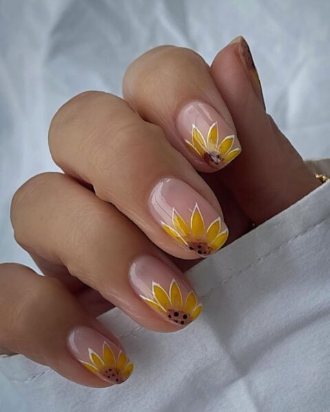 Zdjęcie dłoni ze zbliżeniem na średniej długości, kwadratowe paznokcie w kolorze nude i ozdobione wzorem słoneczników w stylu corner nail art. Dłoń pozuje na tle białego materiału.