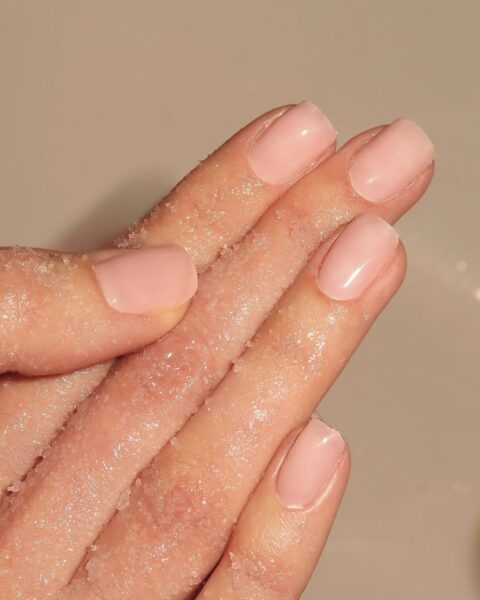 Zdjęcie dłoni ze zbliżeniem na krótkie, kwadratowe paznokcie w kolorze pink nude z połyskiem. Dłonie wykonują zabieg peelingu.