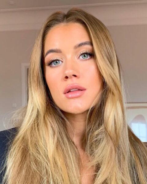 Zdjęcie portretowe kobiety ze zbliżeniem na jej twarz i soft-sculpted make-up, czyli z delikatnym konturowaniem. Ma bardzo długie blond włosy rozpuszczone na ramiona. Ubrana jest w granatową marynarkę. Pozuje w pokoju, w tle widać komodę z plakatem w antyramie.