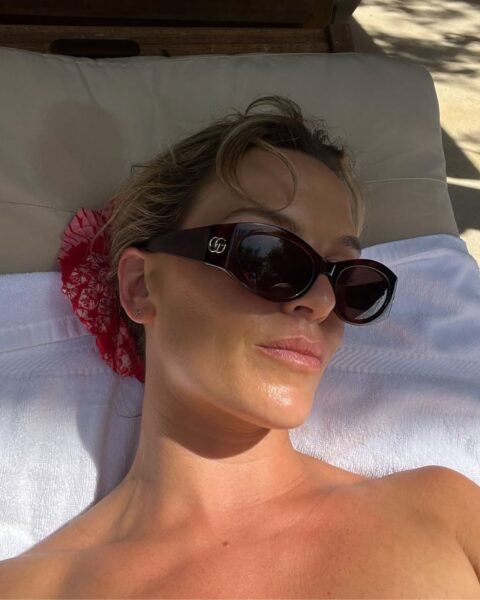 Zdjęcie kobiety leżącej na leżaku plażowym. Zbliżenie na jej twarz i odsłoniety dekolt. Ma blond włosy związane czerwonym scrunchie, a na nosie okulary przeciwsłoneczne. Kobieta chroni się przed słońcem, by uniknąć oparzeń słonecznych.