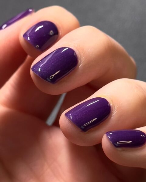 Zdjęcie dłoni ze zbliżeniem na krótkie, owalne paznokcie pomalowane na kolor ciemnego fioletu. To prosty, klasyczny manicure.