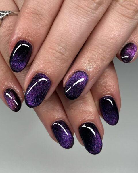 Zdjęcie dłoni ze zbliżeniem na krótkie, owalne paznokcie pomalowane na kolor fioletowy i ozdobione efektem cat eye. Dłonie układają się tak, by prezentować manicure.