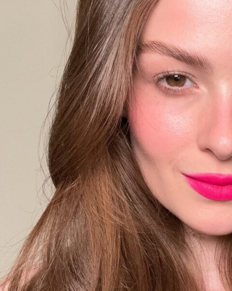 Zdjęcie ze zbliżeniem na twarzy kobiety, która testuje w aplikacji mobilnej makijaż, bo sięga po trend cyber beauty. Ma ciemne, rozpuszczone włosy, wytuszowane rzęsy, dużo rożu na policzkach i różowe usta.