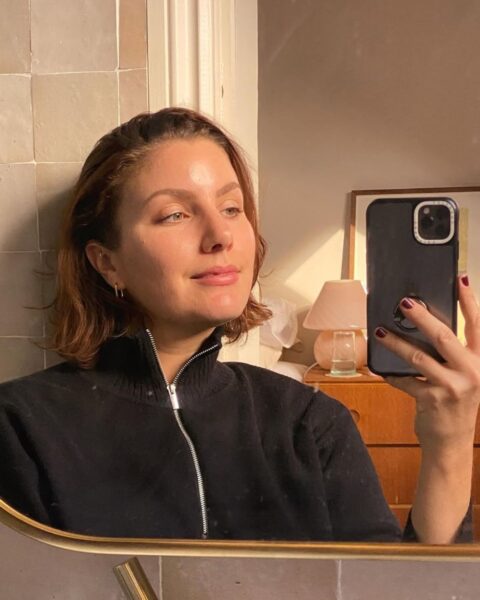 Zdjęcie kobiety, która zrobiła sobie selfie w lustrze ze złotą ramką. Lustro wisi w łazience, na szarych kafelkach. Kobieta zaś ma czarną bluzę z zamkiem błyskawicznym i stójką. Ma ciemne włosy do ramion.