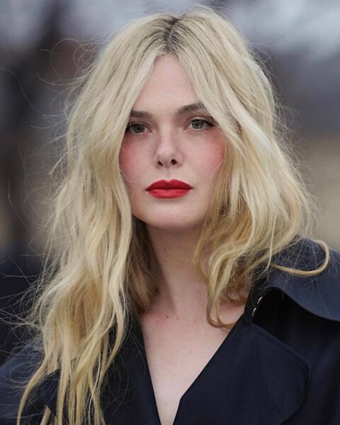 Zdjęcie aktorki Elle Fanning, która ma włosy lemon platinum blonde. Włosy są długie, falowane, lekko zmierzwione i opadają na ramiona. Aktorka jest ubrana w czarny trencz.
