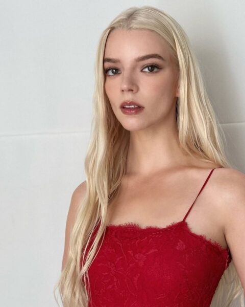Zdjęcie aktorki Anyi Taylor Joy, która ma długie blond włosy w odcieniu cytrynowej platyny. Ubrana jest w czerwony top na ramiączkach. Pozuje na tle białej ściany.