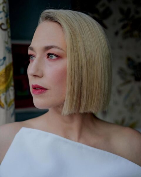 Zdjęcie aktorki Carrie Coon w krótkim blond bobie o odcieniu cytrynowej platyny. Włosy sa idealnie wyprostowane i ścięte równo, jak blunt bob. Kobieta patrzy w bok. Ma na sobie biały top.