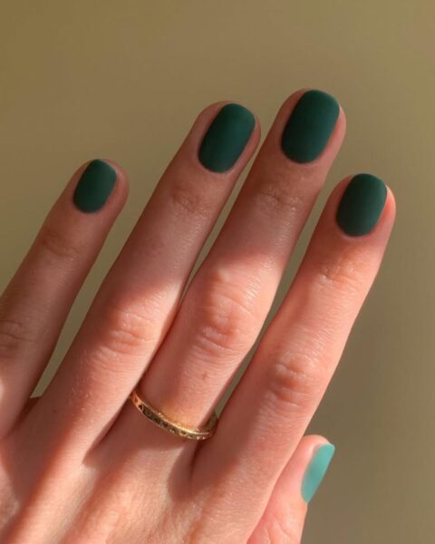 Zdjęcie dłoni z krótkimi, owalnymi paznokciami w kolorze zielonym z efektem suede nails. Na środkowym palcu znajduje się złota, cienka obrączka.