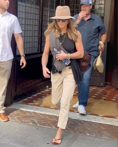 Kobieta w bezowym kapeluszu, spodniach 3/4, ciemnym topie. Na ramieniu ma dużą torebkę - Jennifer Aniston.