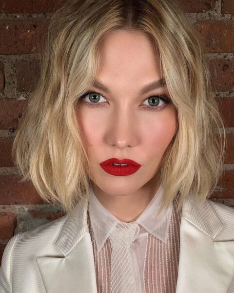 Zdjęcie kobiety z fryzurą boho bob w kolorze blond i lekkimi falami. Jest ubrana w białą koszulę półtransparentną oraz białą marynarką. Pozuje na tle ceglanej ściany. Ma usta pomalowane na czerwono.