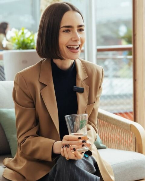 Uśmiechnięta Lily Collins siedzi na kanapie w beżowej marynarce, czarnym golfie i szarych spodniach w paski, trzymając w ręku szklankę z wodą. Włosy ma ułożone w krótkiego, prostego boba, a na klapie marynarki przypięty jest mały mikrofon. Na pierwszym planie widać puszki z napojami.