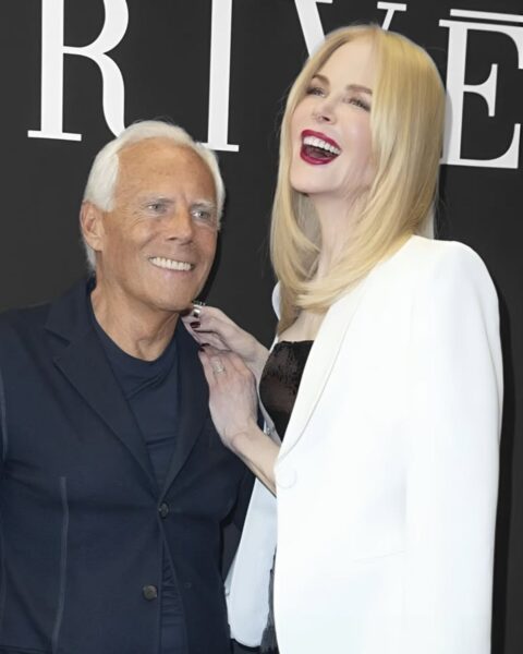 Giorgio Armani i Nicole Kidman pozują razem na tle czarnego baneru z napisem "PRIVÉ". Armani ubrany jest w granatową marynarkę i koszulkę, uśmiecha się, patrząc w bok. Nicole Kidman, z uśmiechem i uniesioną głową, ubrana jest w białą marynarkę i czarną, błyszczącą sukienkę, a jej dłoń spoczywa na ramieniu projektanta.